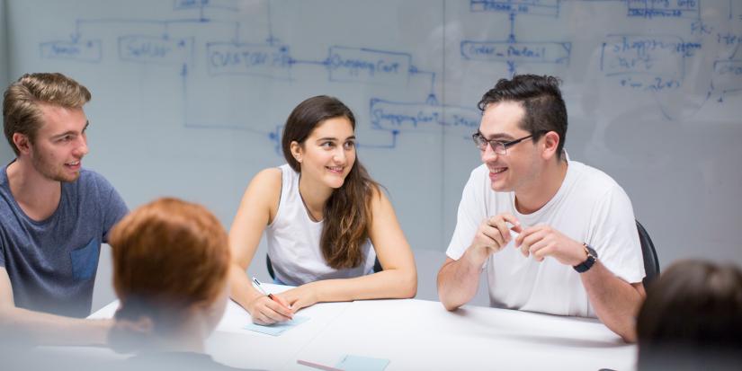 Five students in the Software development studio, sitting at a table and smiling. Behind them is a whiteboard wall