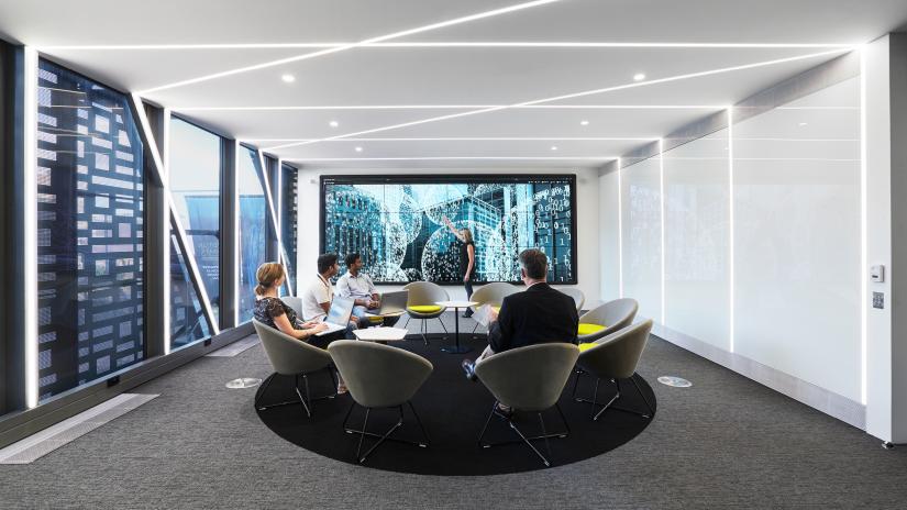 Photo of the UTS DataLounge. Four people are sitting in a circle of chairs looking towards the LCD screen where another person is presenting.