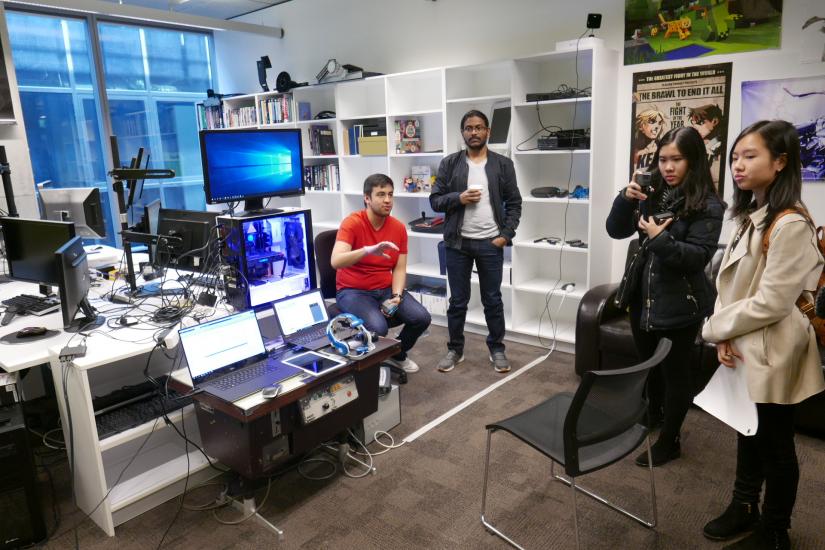 Four people sit and stand in a brightly lit room full of computer screens and gadgets on shelves, and look to the left of the camera