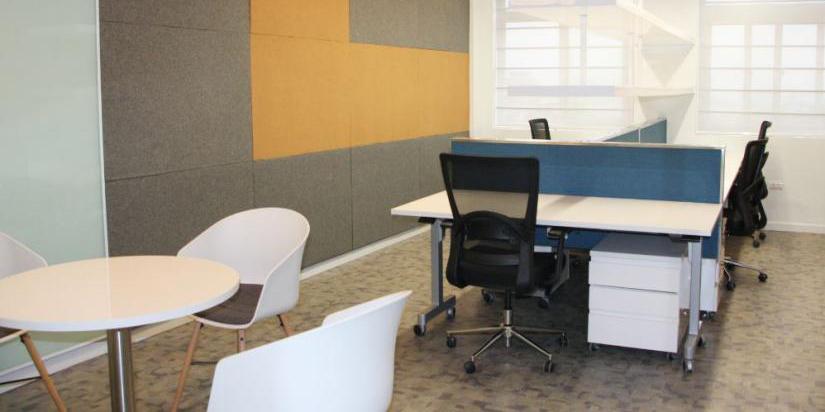 A brightly lit room features open plan seating and desks and a wall with a bright yellow panel