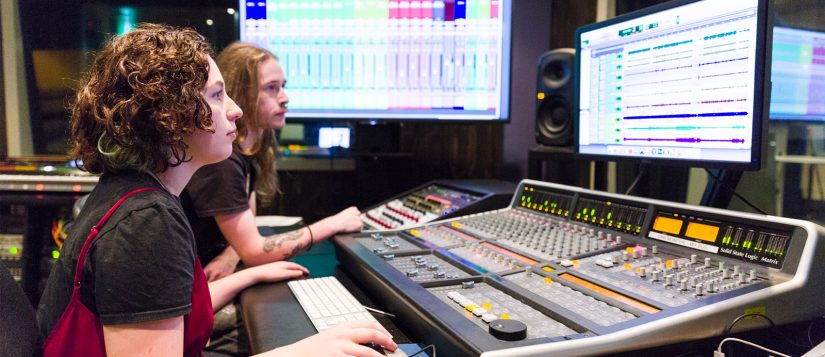 Two students sitting in a sound recording booth, looking at levels