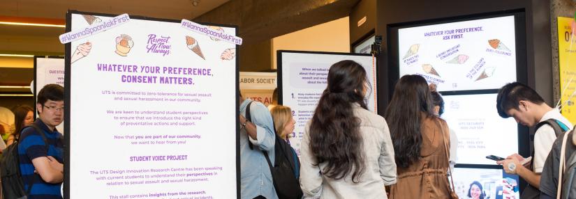 A group of people scattered between a tall board and screens. The board says 'Whatever your preference, consent matters'
