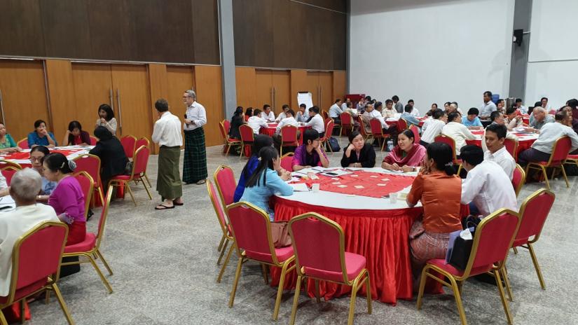 A room full of people sit around red draped tables