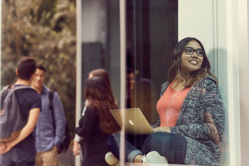 A smiling student, sitting with a laptop whilst looking out the window to a group of students