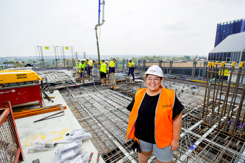 Young woman in hard hat and orange vest stands leading a team of men on a construction site