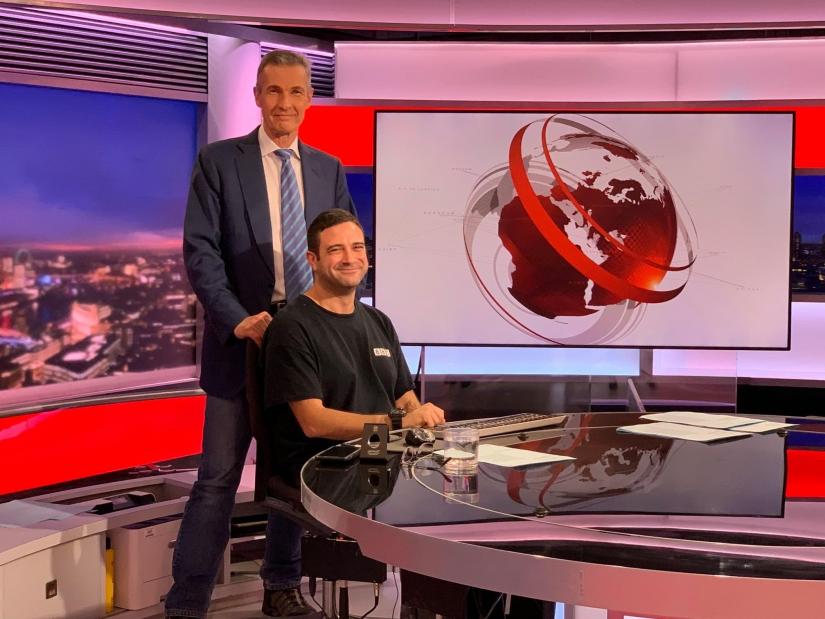 Seref sitting at the news desk of BBC World with journalist Mike Embley standing behind him