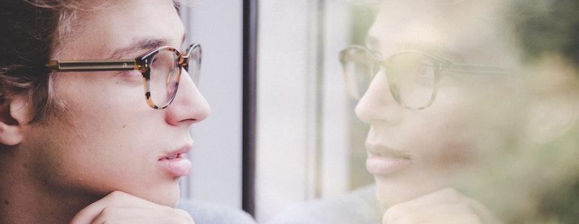 Young man with glasses looking at reflection in window