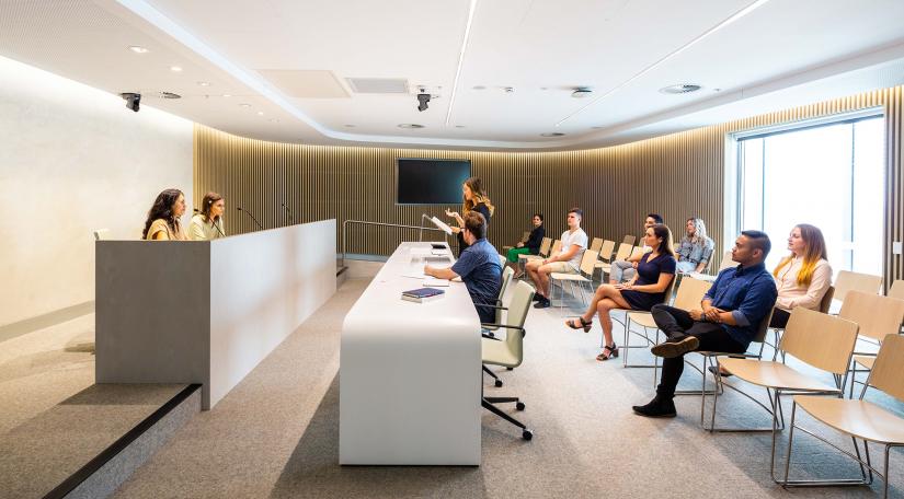 Student 'judges' sit at a high table. Below them, student-advocates sit behind a desk or stand at a lectern presenting an argument. More students sit in chairs watching on.
