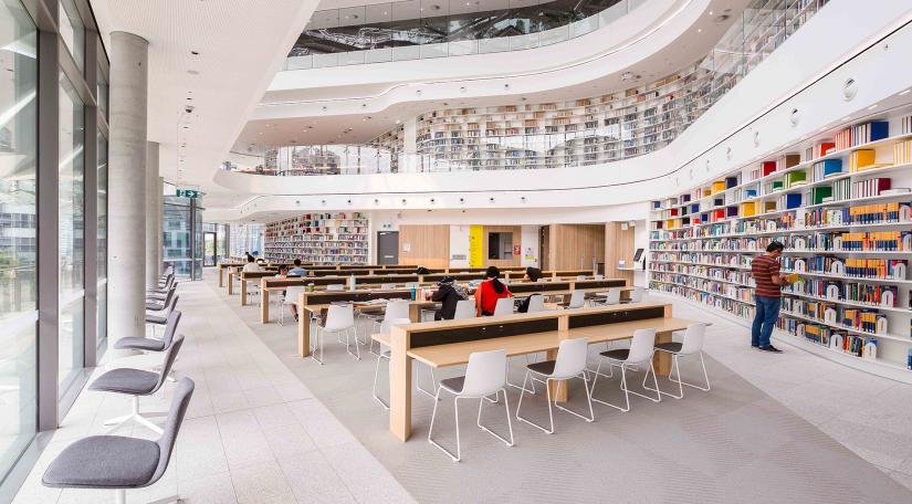 A large, high-ceilinged room with curved walls covered in white bookshelves filled with colourful books, with traditional wooden desks and large windows to outside.