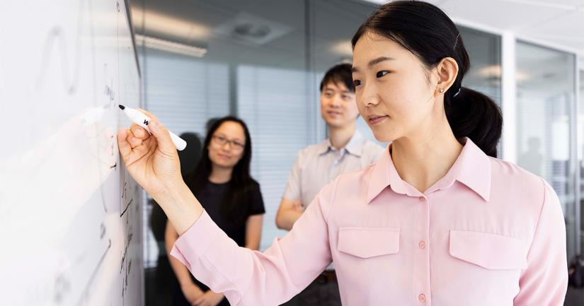 Young female AAII researcher in pink shirt and hair tied back writes on a whiteboard while a female and male researcher are watching in the background