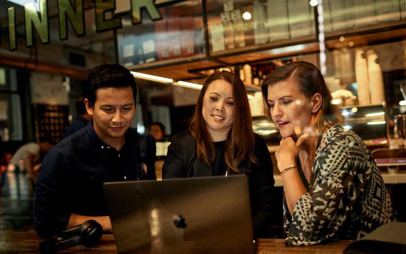 A man and two women sit at a cafe table and look at a laptop screen.