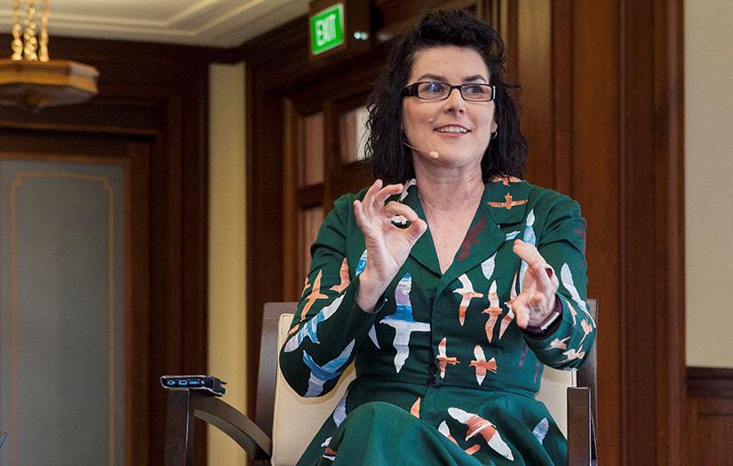 Woman seated in a green dress with short dark hair wearing glasses and gesturing with her hands 