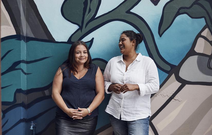 Two young woman standing in front of a colourful artwork smiling and laughing.