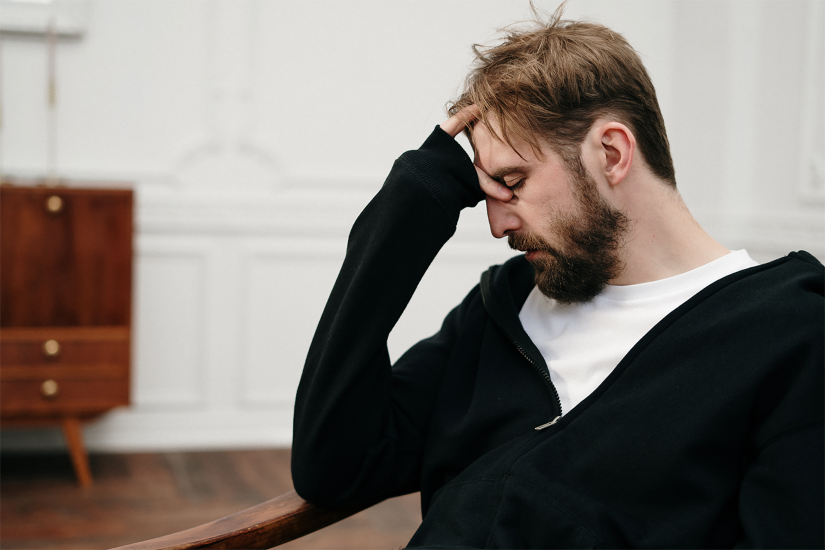 A man with short hair and a beard sits with his head resting on his hand. He is wearing a white t-shirt under a black jumper.