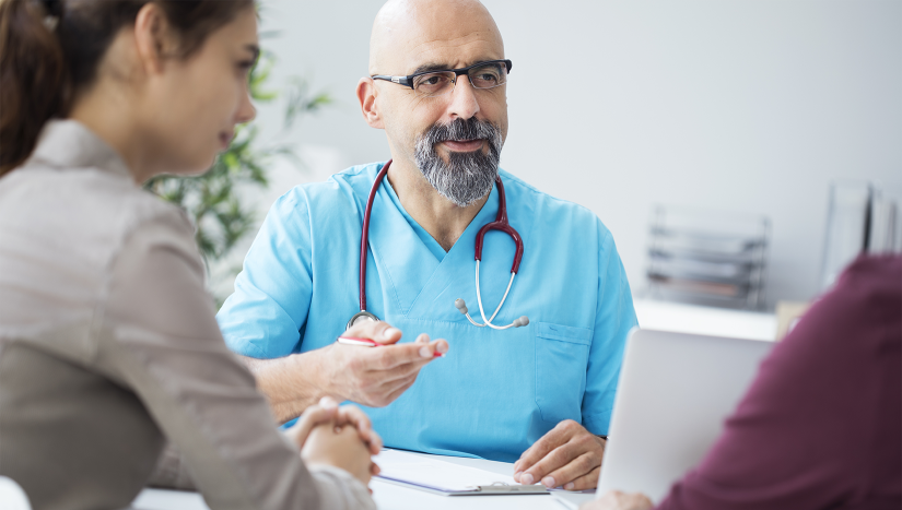 A doctor with a bald head and a beard is wearing blue scrubs and a stethoscope around his neck. He is sitting at a desk talking to two people, one in a red shirt and one in a grey shirt with brown hair in a ponytail.