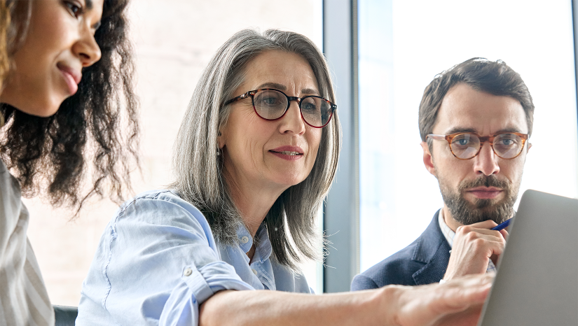 Mature woman with shoulder-length grey hair and glasses is pointing at a computer screen. Either side of her sit two younger team members - a man with brown hair and a beard, wearing glasses and a woman with long, brown curly hair.