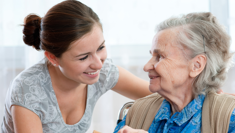 elderly lady with grey hair wearing blue shirt and tan vest is smiling at her carer, a young woman with brown ahid in a bu wearing a short-sleeved grey top.