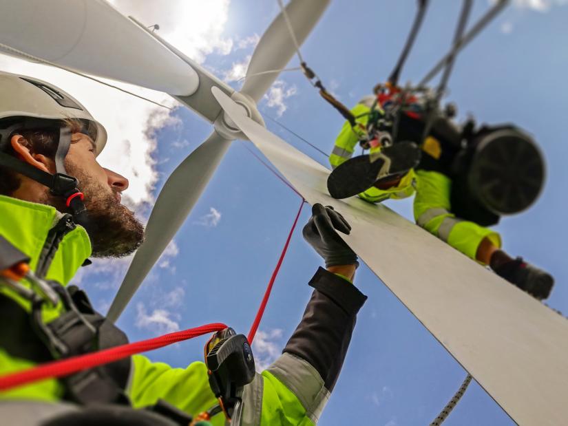 An energy worker in yellow hi-vis gear stands at the base of a wind turbine. He is looking up at another worker who is on scaffolding.