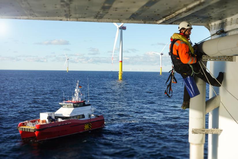 A man in a harness and hi-vis gear is working on the underside of a concrete structure. There is a ship on the water behind him.