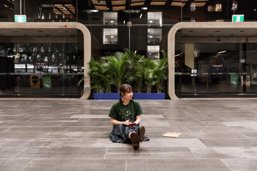 A person sits on the ground between two large doors. They look to their left and hold a microphone in their hands. 