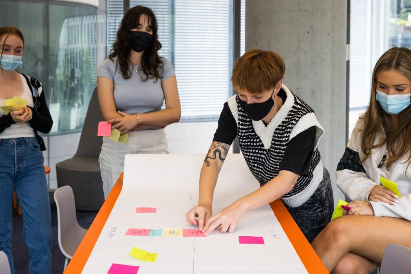 People stand beside a table with a large piece of paper. They have placed post it noteson the paper. 
