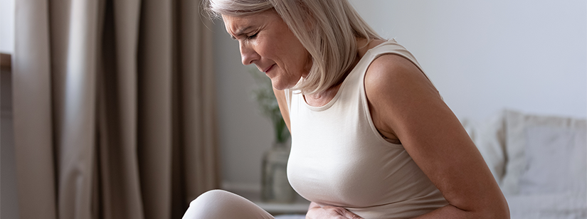 Woman wearing beige top and trousers sits on bed clutching her abdomen