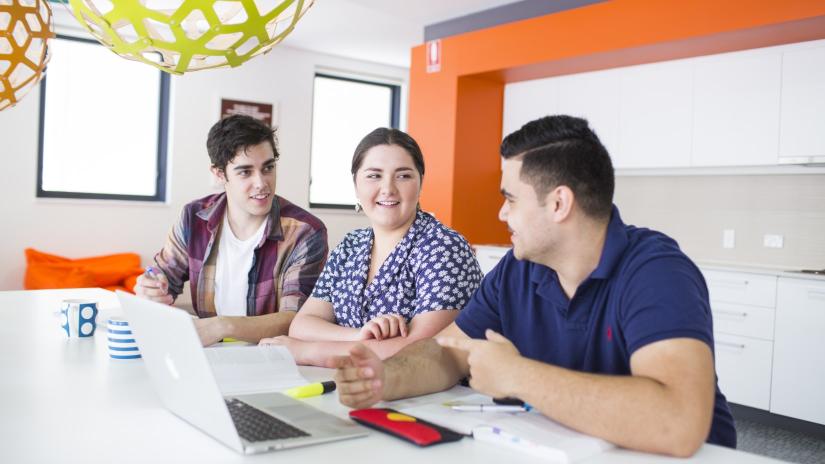 Three Indigenous students are working at a table and engaging in casual conversation.