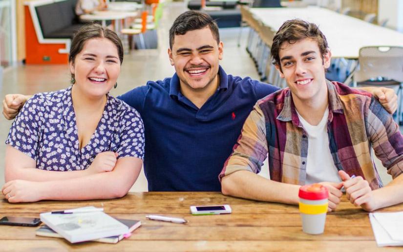 Three students sitting at a desk with papers and a coffee cup, smiling.