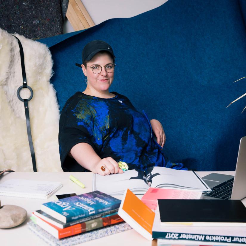 A smiling person sits against a blue backdrop. The table in front of them has stacks of books and an open laptop computer on it. 