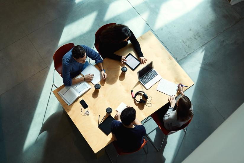 An aerial photo of four people working around a wooden table which has textbooks, laptops and take away coffee cups on it