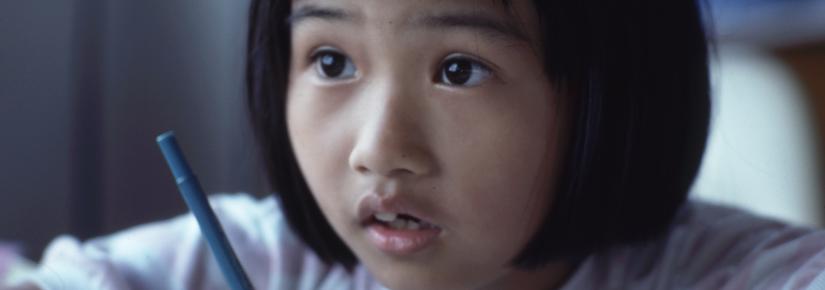 A student looks attentively towards the front of the classroom with pencil poised to write.