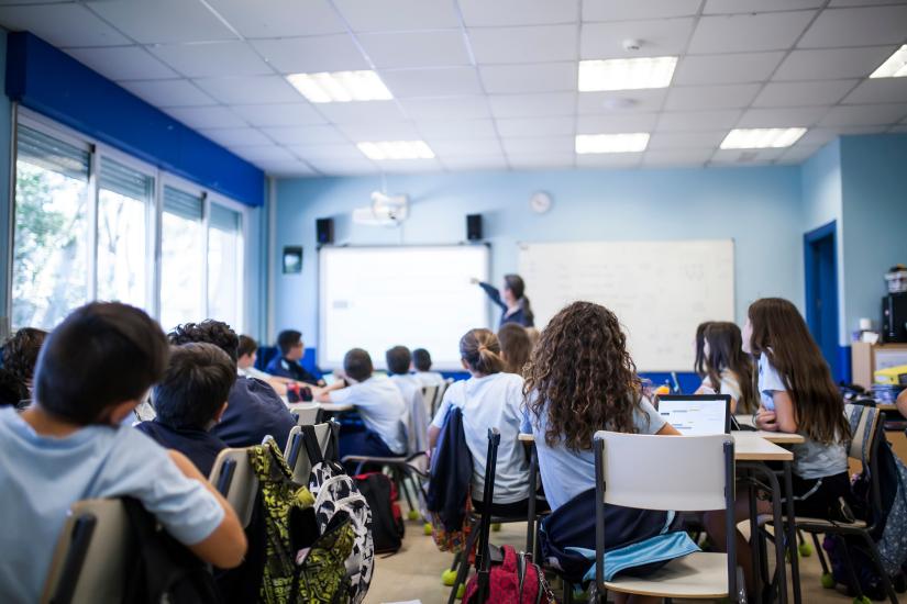 View of a high school classroom, students watch as a teacher writes on whiteboard