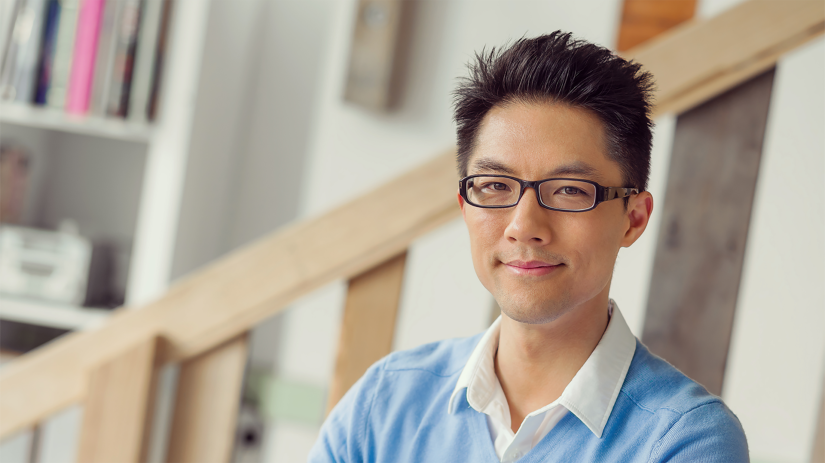 Young Asian man in glasses, white collared shirt and blue jumper sitting at a desk with a white laptop