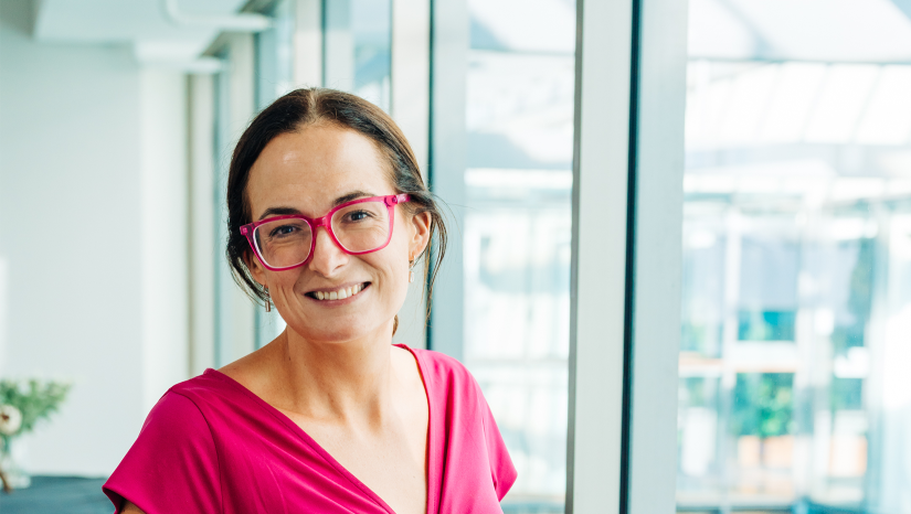 Dr Caitlin Sheehan has dark hair in pony tail. She is wearing pink-framed glasses and a pink dress. She is smiling at the camera. 