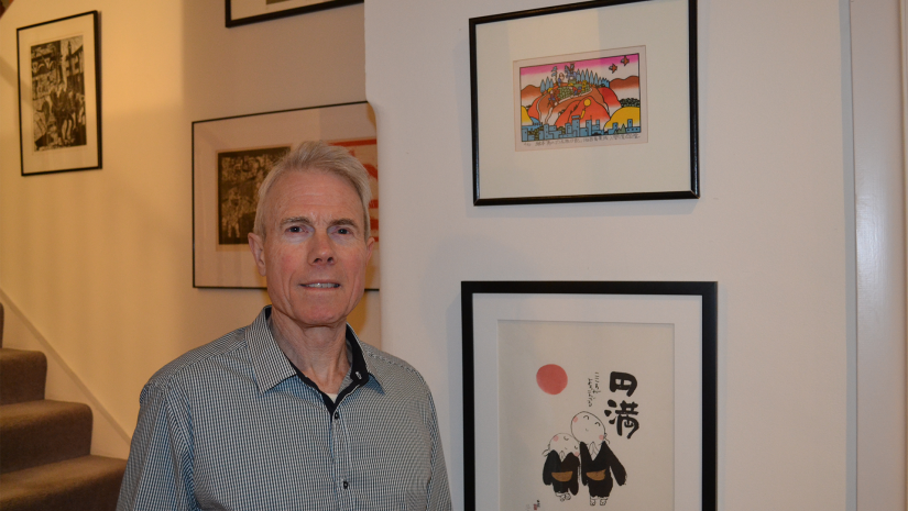 Stephen Peterson has short grey hair and he is wearing a button up shirt. He is standing in front of a gallery wall of paintings.