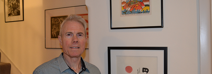 Stephen Peterson has short grey hair and he is wearing a button up shirt. He is standing in front of a gallery wall of paintings.