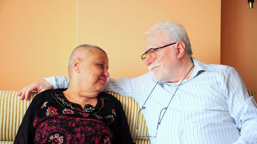 Lady and her husband sitting on a couch side by side smiling at each other. The lad is wearing a long sleeved black top with a red pattern on the front. She has lost her hair due to cancer treatment. The main is wearing a striped button up shirt. He has grey hair and glasses.