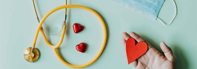 Birds eye view of a hand holding a paper heart, a stethosope, face mask and heart-shaped chocolates.