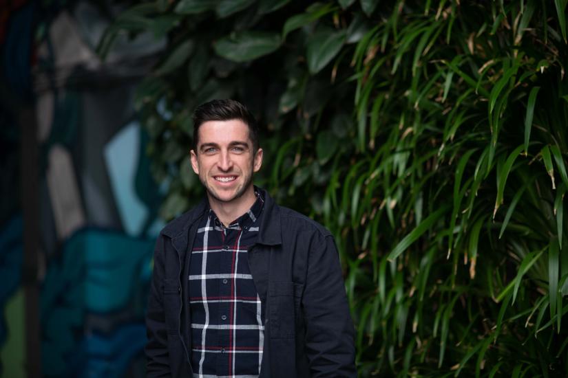 A man stands smiling at the camera, a green wall in the background