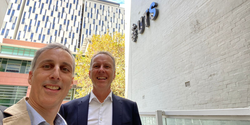 Professors Enzo Sauma and Damien Giurco stand left of frame next to a brick wall painted white with black UTS text logo signage. Yellowed tree leaves can be seen behind them.