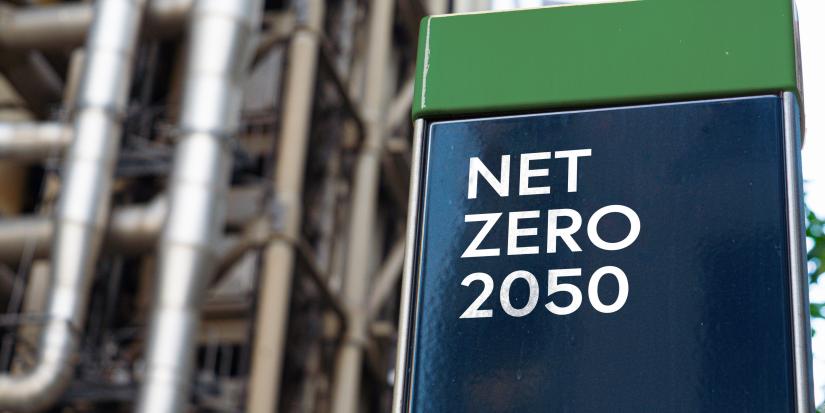 A blue and green shiny block says "NET ZERO 2050" in white capital letters. Pipes and infrastructure can be seen blurry in the background.