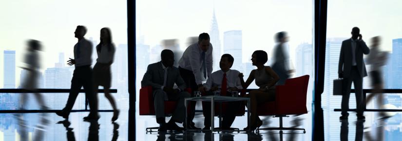 a group of people gathered in a large office around a table with large windows in the background.