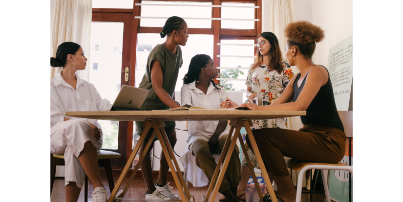 Five women of varying cultural backgrounds are gathered around a table, one of the women is addressing the group.