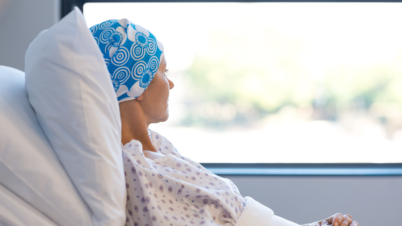 Young woman in hospital bed leaning against pillows in white pillow cases. She is wearing a floral nightie and has a blue and white scarf on her head. 