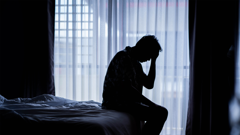 Silhouette of a man sitting on a bed with his hand held to his head. There are net curtains on a window in the background. 