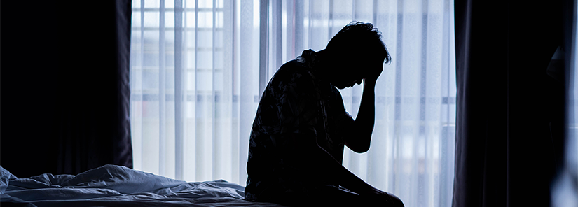 Silhouette of a man sitting on a bed with his hand held to his head. There are net curtains on a window in the background. 