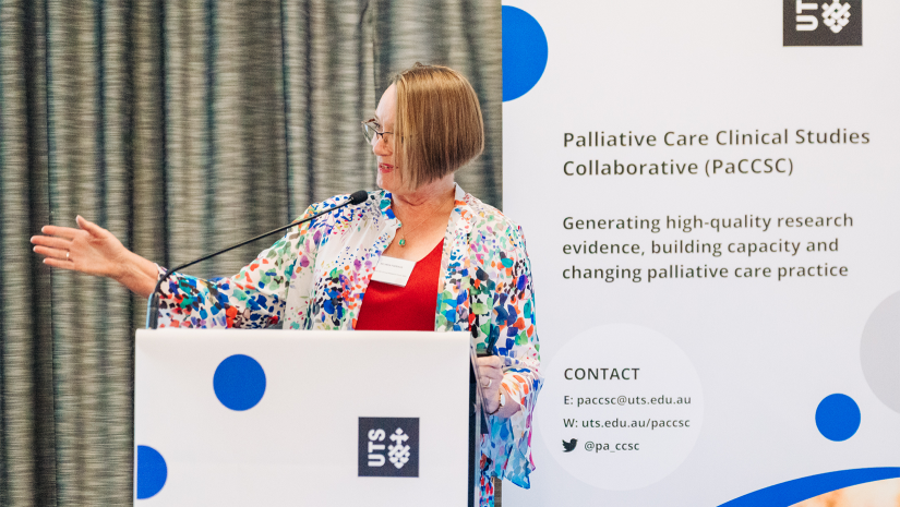 Belinda Fazekas presenting at a lectern. she is standing in front of a PaCCSC promotional banner, which has a grey background and blue spots. Belinda is wearing a red top under a floral cardigan.