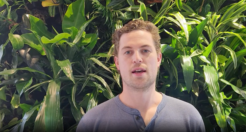 Rupert Legg stands in front of a green wall of plants. He his talking to the camera.