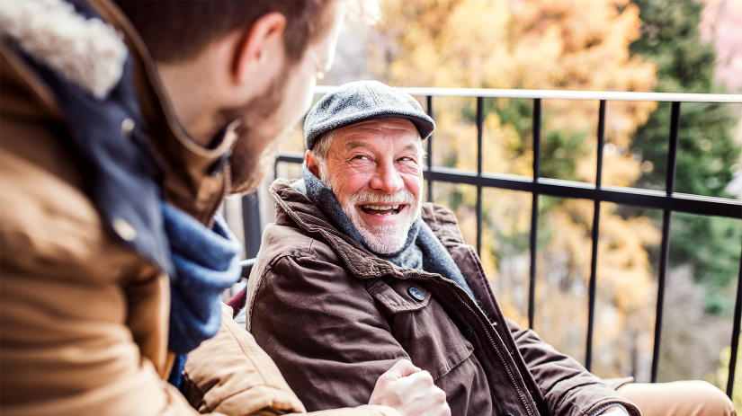 Father and son, both with beards, on a walk. The father is wearing a flat cap, scarf and coat. The son is wearing a scarf and coat. The are looking at each other and smiling.