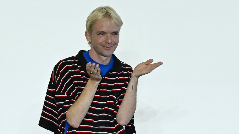 Alix Higgins stands in front of a white wall with slight smile and hands up in front of face